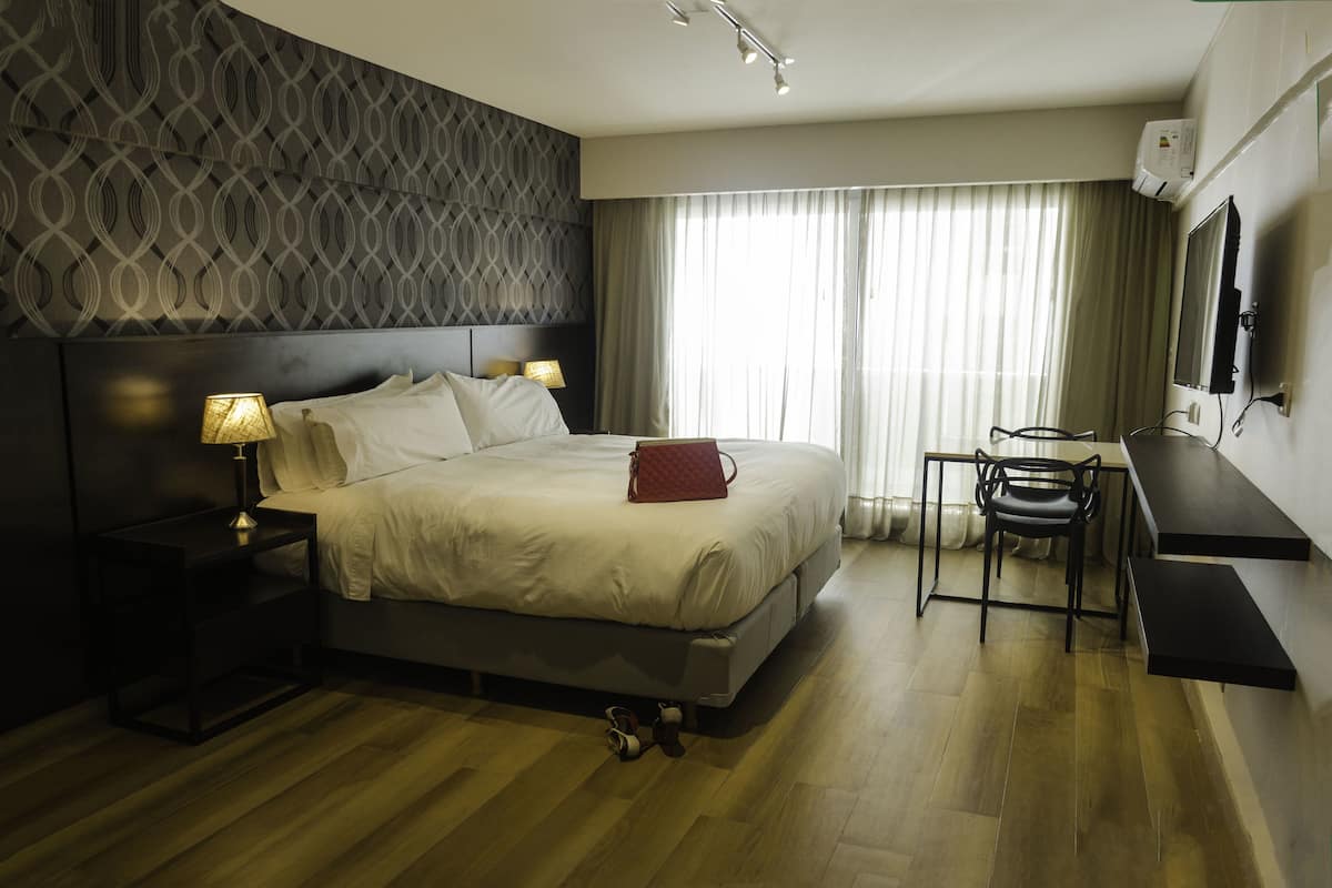 A modern hotel room with a large bed, bedside lamps, a desk with a chair, and a large window with curtains.
