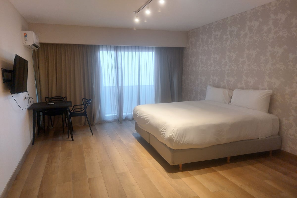 A hotel room with a large bed, a desk with a chair, a TV mounted on the wall, and a window with curtains.