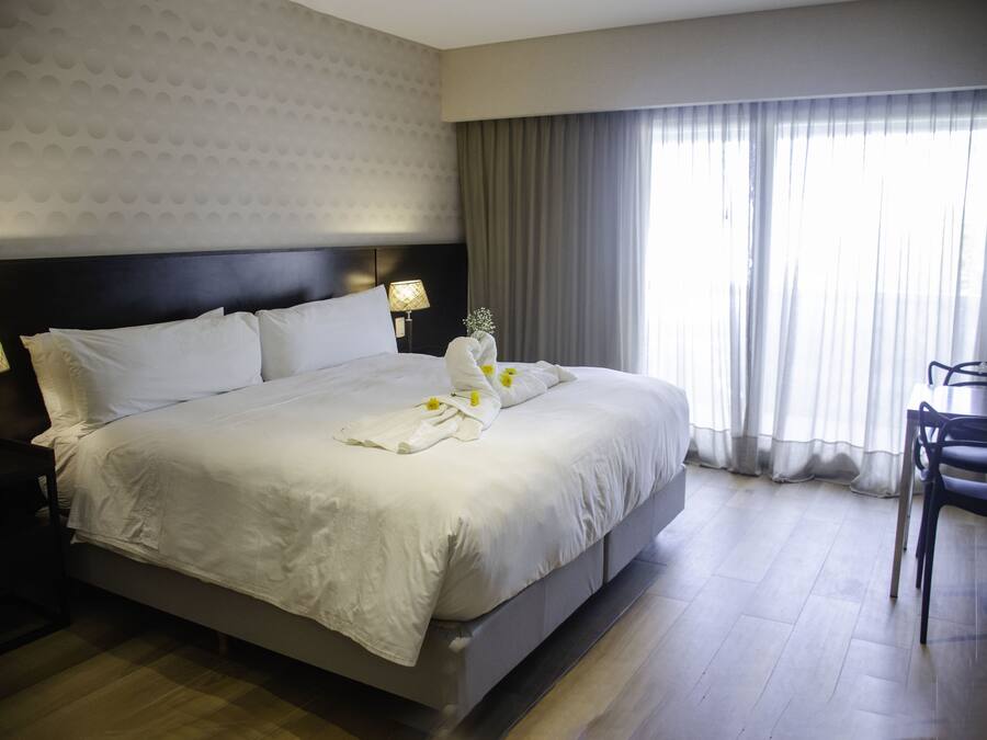 A hotel room with a large bed, a bedside table with a lamp, a dining area with a table and chairs, and a window with curtains.
