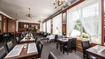 A hotel dining area with multiple tables and chairs, large windows with sheer curtains, and a chandelier.