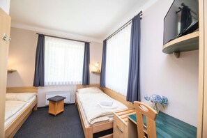 Habitación de hotel con dos camas individuales, una mesita pequeña, una silla, un televisor montado en la pared y una ventana con cortinas.