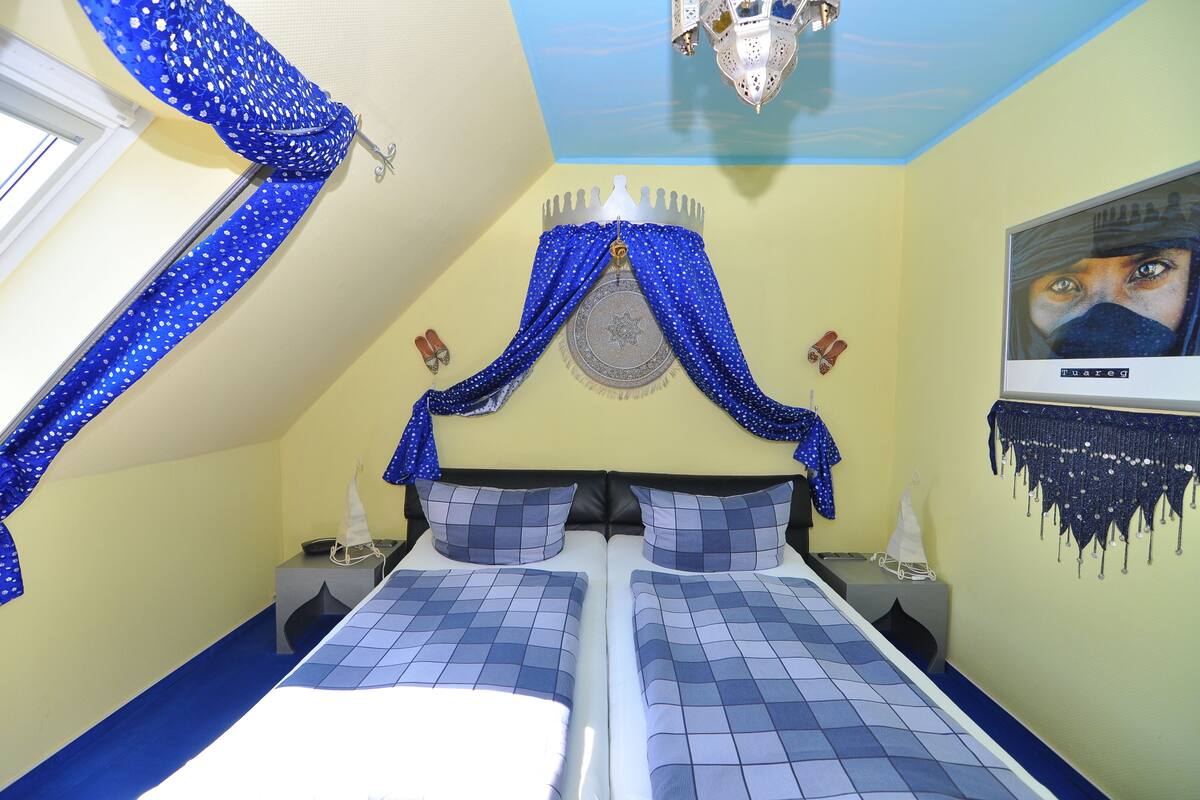 Une chambre avec deux lits, un lustre, une fenêtre avec des rideaux bleus et un élément décoratif mural.