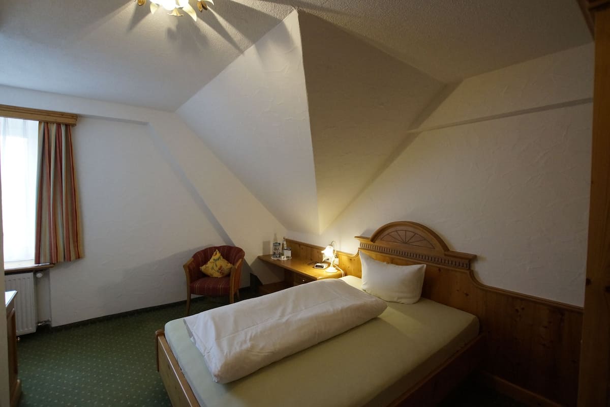 Una habitación pequeña y acogedora con una cama individual, cabecera de madera, escritorio con lámpara y una silla.