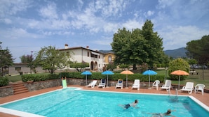 Ein Schwimmbecken mit Liegestühlen und Sonnenschirmen, umgeben von einem angelegten Garten und einem Gebäude im Hintergrund.