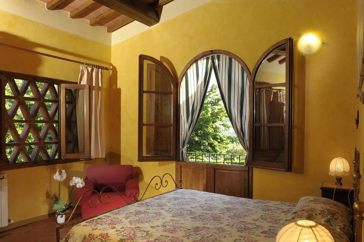 A bedroom with a large bed, wooden beams, a window with curtains, and a nightstand with a lamp.