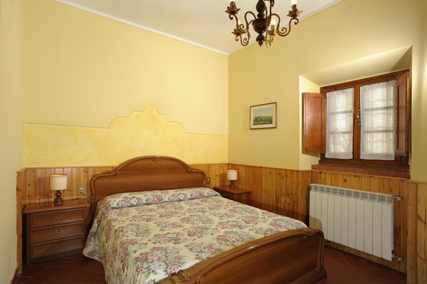 A bedroom with a wooden bed, a nightstand, a lamp, a radiator, and a window with curtains. - Agriturismo Bellosguardo (Reggello)