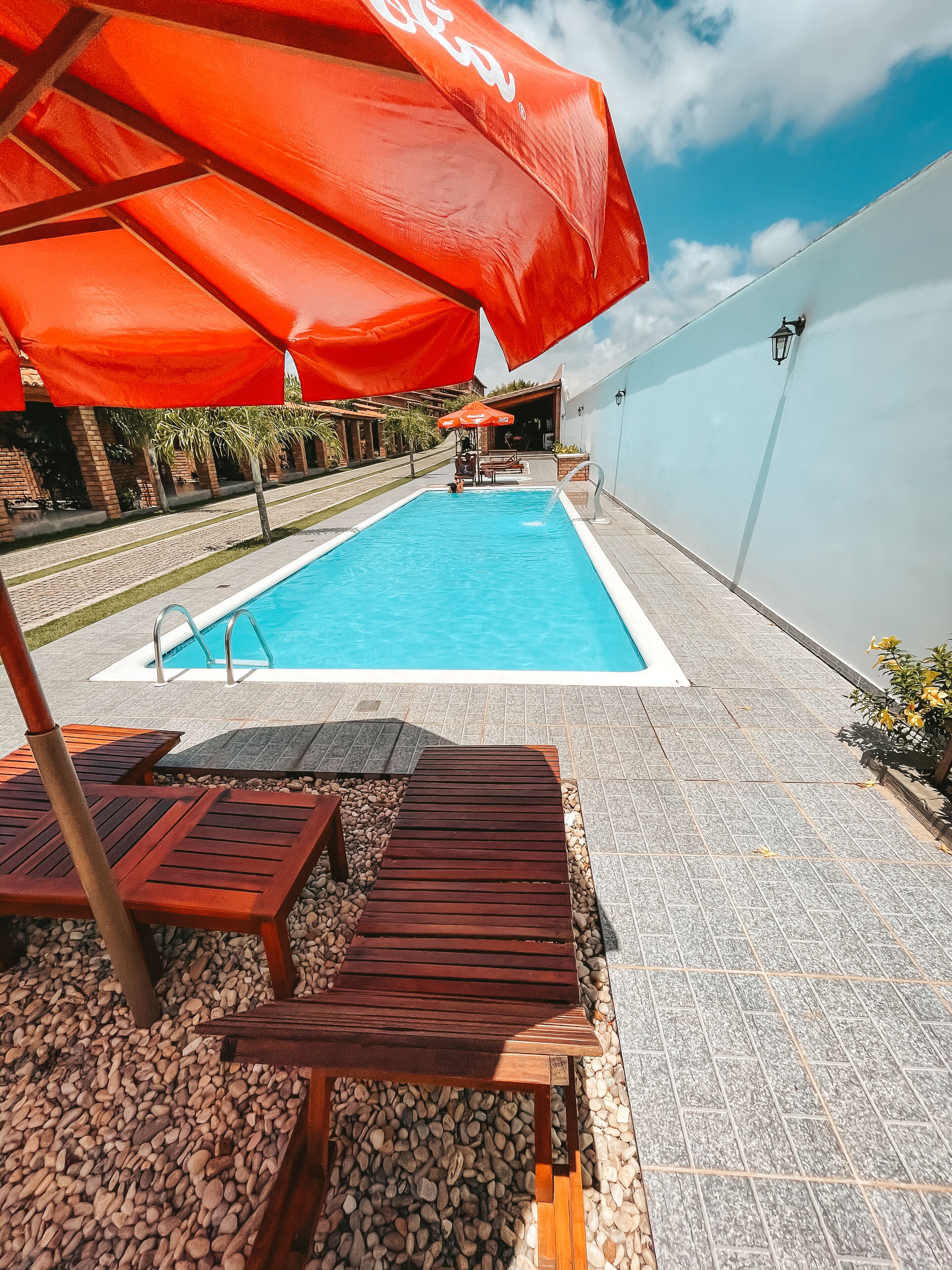 A swimming pool with a tiled deck, wooden lounge chairs, and red umbrellas.