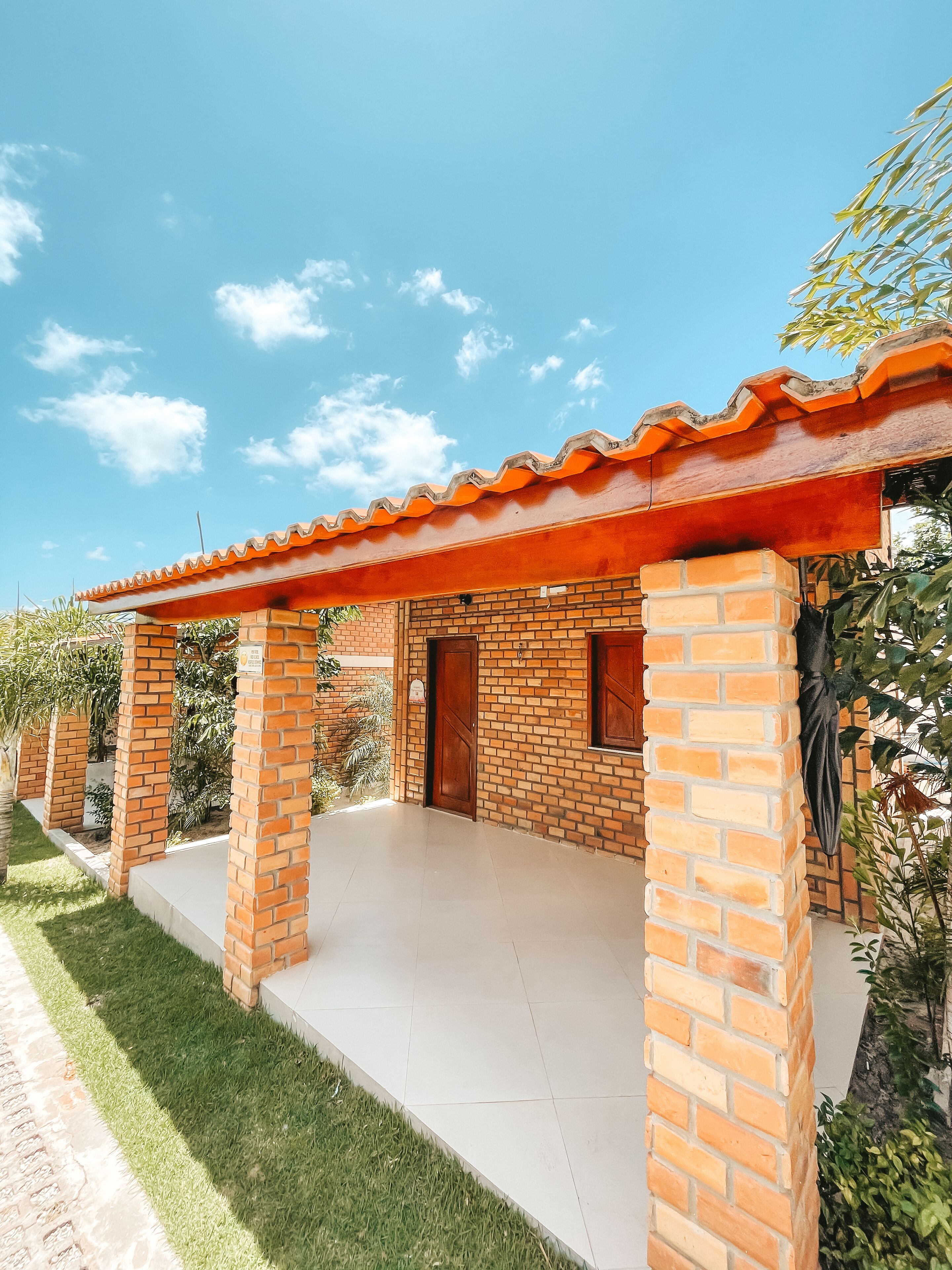 A covered outdoor area with brick pillars and a tiled floor, surrounded by greenery.