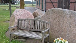A wooden bench with a metal backrest, positioned on grass with large rocks nearby and a potted plant with flowers to the right. - Landhaus Bondzio (Langen Brütz)