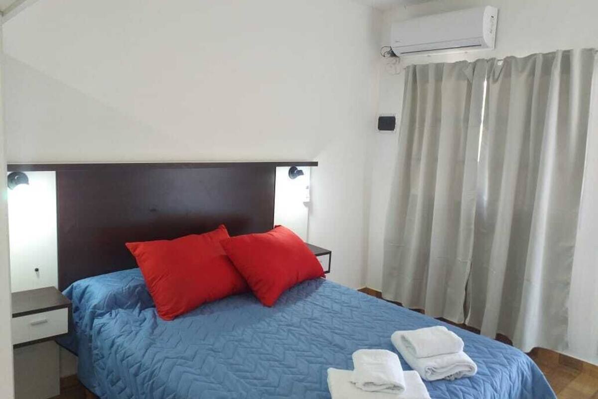 A bedroom with a bed, two red pillows, a blue bedspread, a nightstand, a drawer, and an air conditioning unit.
