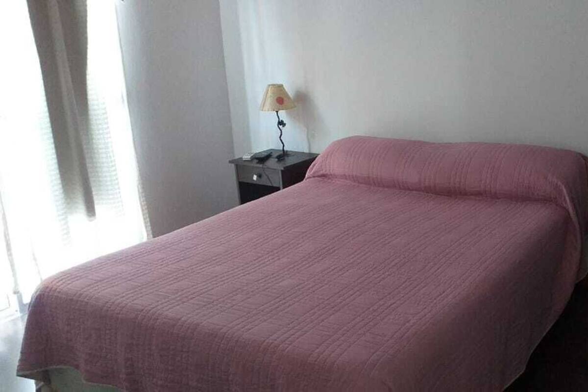 A single bed with a pink bedspread in a room with a small bedside table and a lamp.