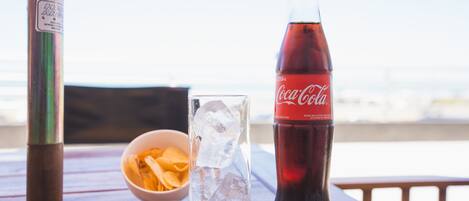 Un vaso de Coca-Cola, un tazón de papas fritas y una botella de Coca-Cola sobre una mesa de madera con vista a la playa.