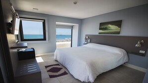 Un dormitorio con una cama grande, un escritorio, una silla, un ventanal con vistas a la playa y un cuadro en la pared.
