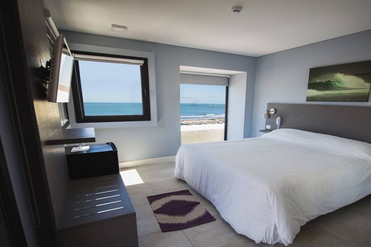 A bedroom with a large bed, a desk, a chair, a window with a view of the beach, and a painting on the wall.