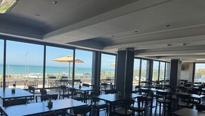 Un comedor amplio con grandes ventanales que ofrecen vistas a la playa y al océano. Mesas y sillas dispuestas para comer.