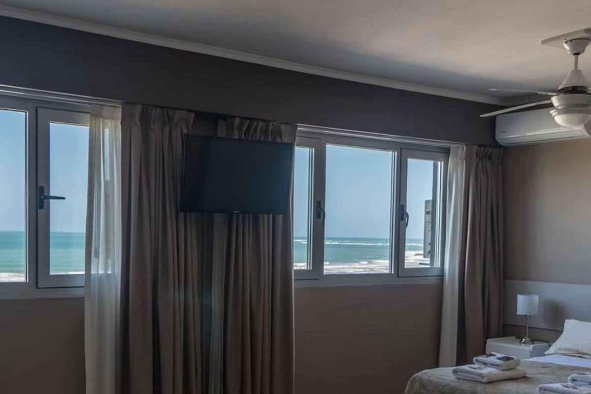 A hotel room with a bed, a television, a ceiling fan, and a large window with a view of the beach.