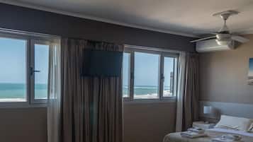 Une chambre d’hôtel équipée d’un lit, d’une télévision, d’un ventilateur de plafond et d’une grande fenêtre donnant sur la plage.