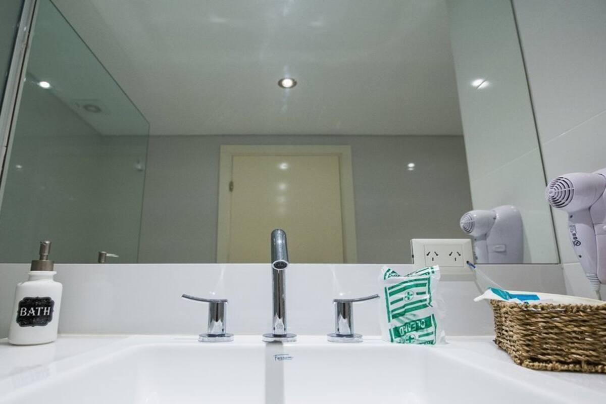 A modern bathroom with a white sink, three faucets, a bottle labeled 'BATH,' a hairdryer, and a basket.
