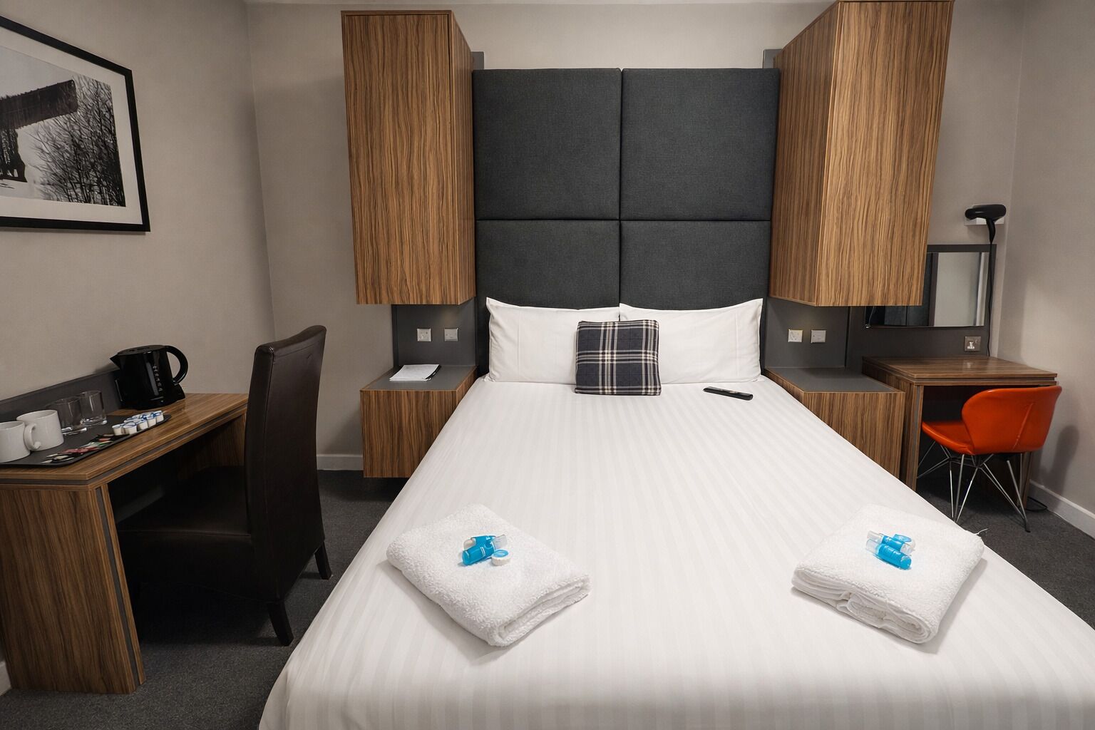 Habitación de hotel con una cama grande, un escritorio con silla, una mesita con otra silla, cafetera y un cuadro en la pared.