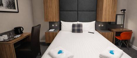 Habitación de hotel con una cama grande, un escritorio con silla, una mesita con otra silla, cafetera y un cuadro en la pared.
