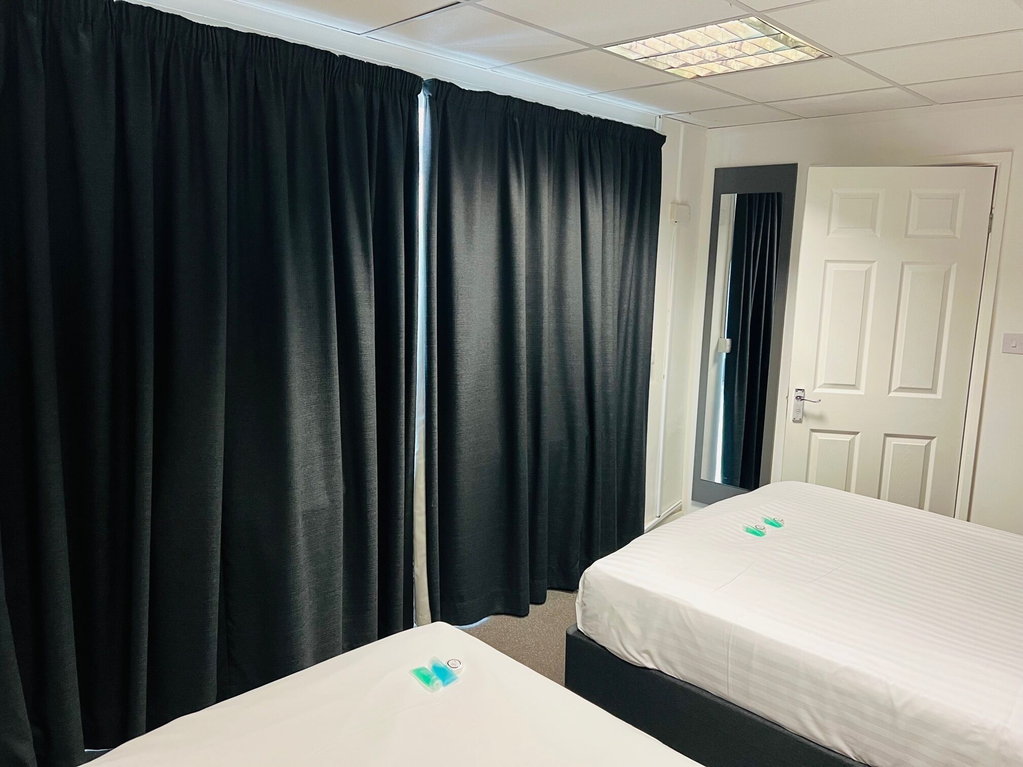 Habitación de hotel con dos camas individuales, cortinas negras, una puerta blanca e iluminación fluorescente en el techo.