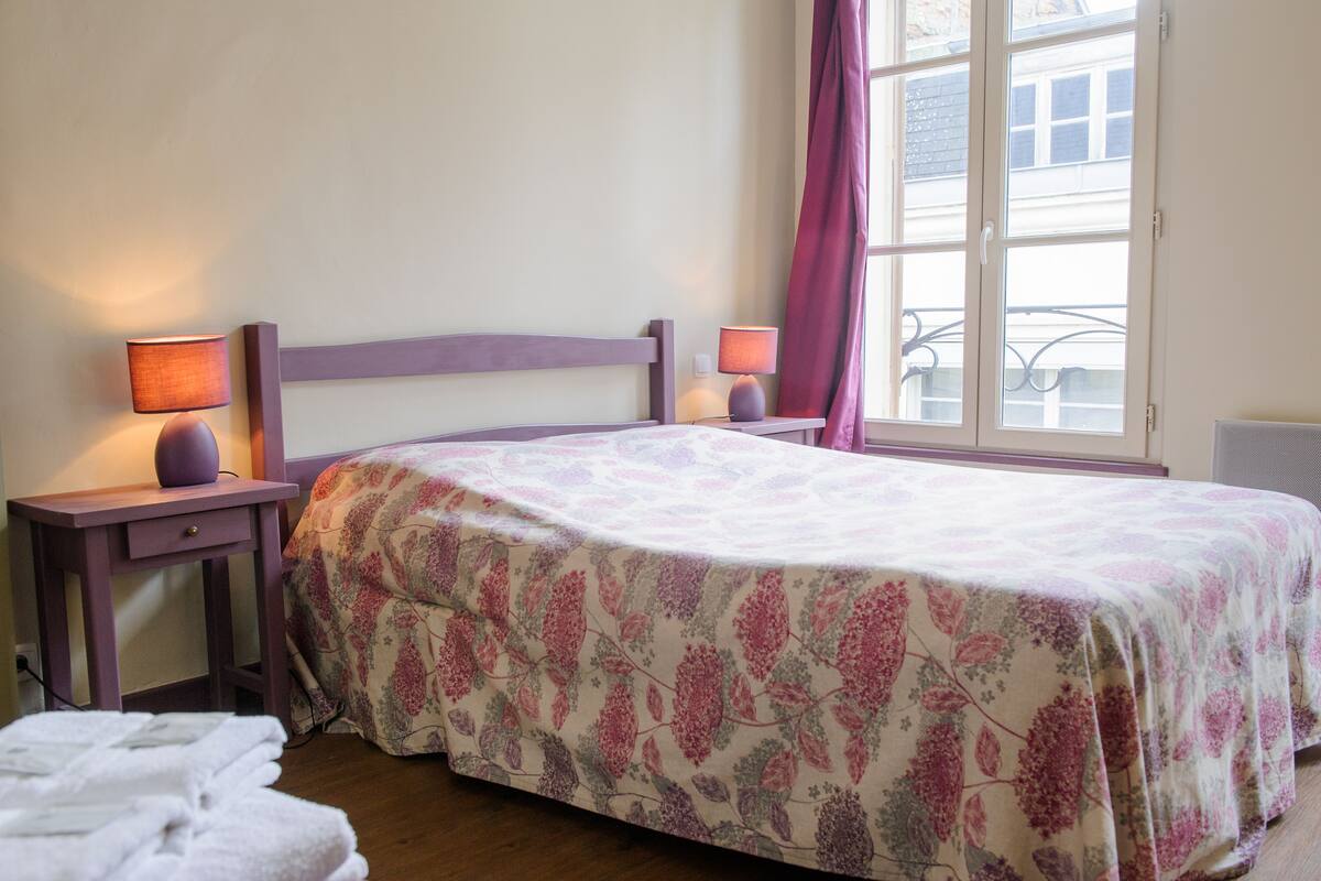A bedroom with a bed, two bedside tables with lamps, a window with curtains, and a radiator.