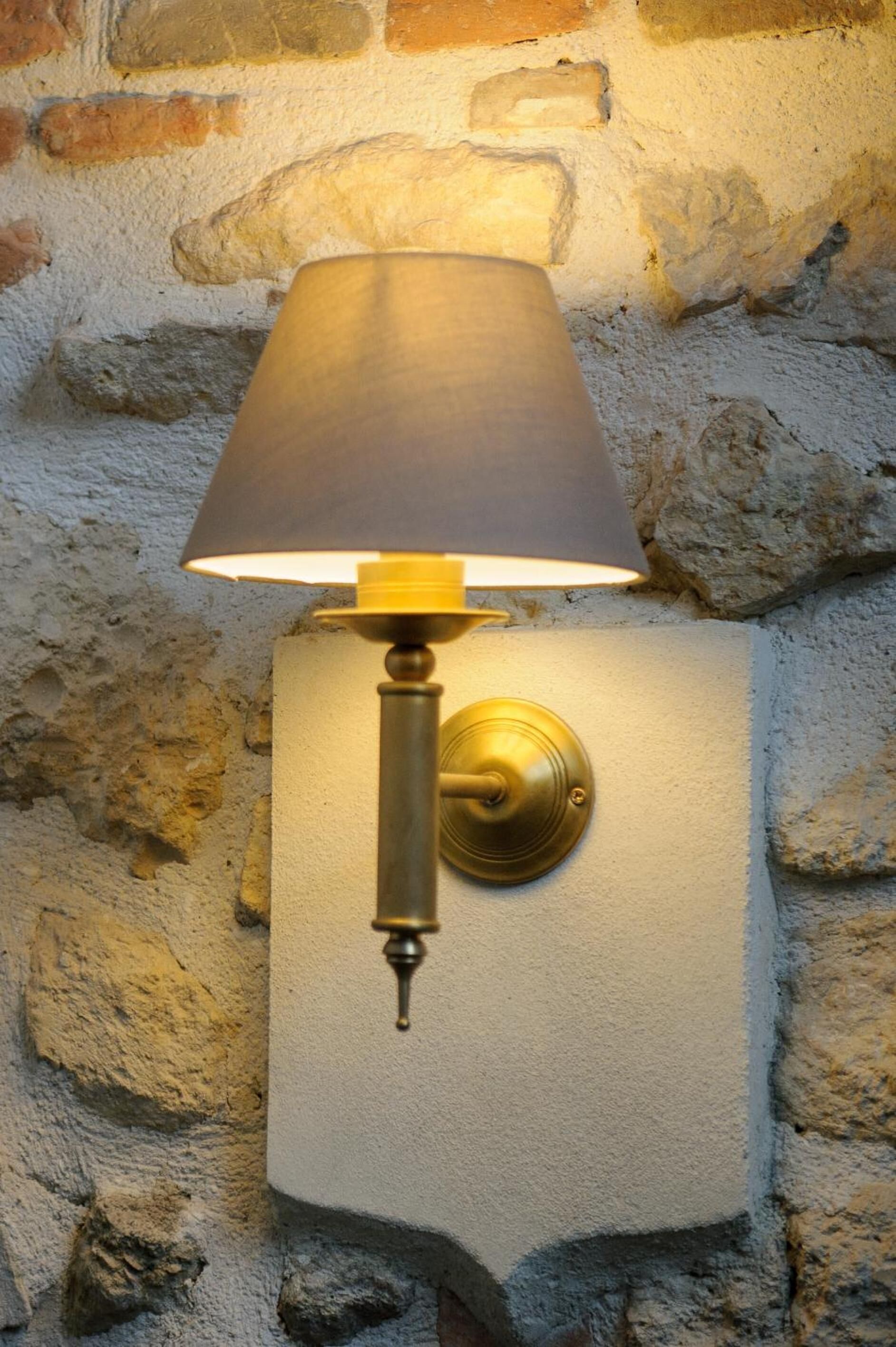 A wall-mounted lamp with a classic design, featuring a brass base and a fabric shade, set against a textured stone wall.