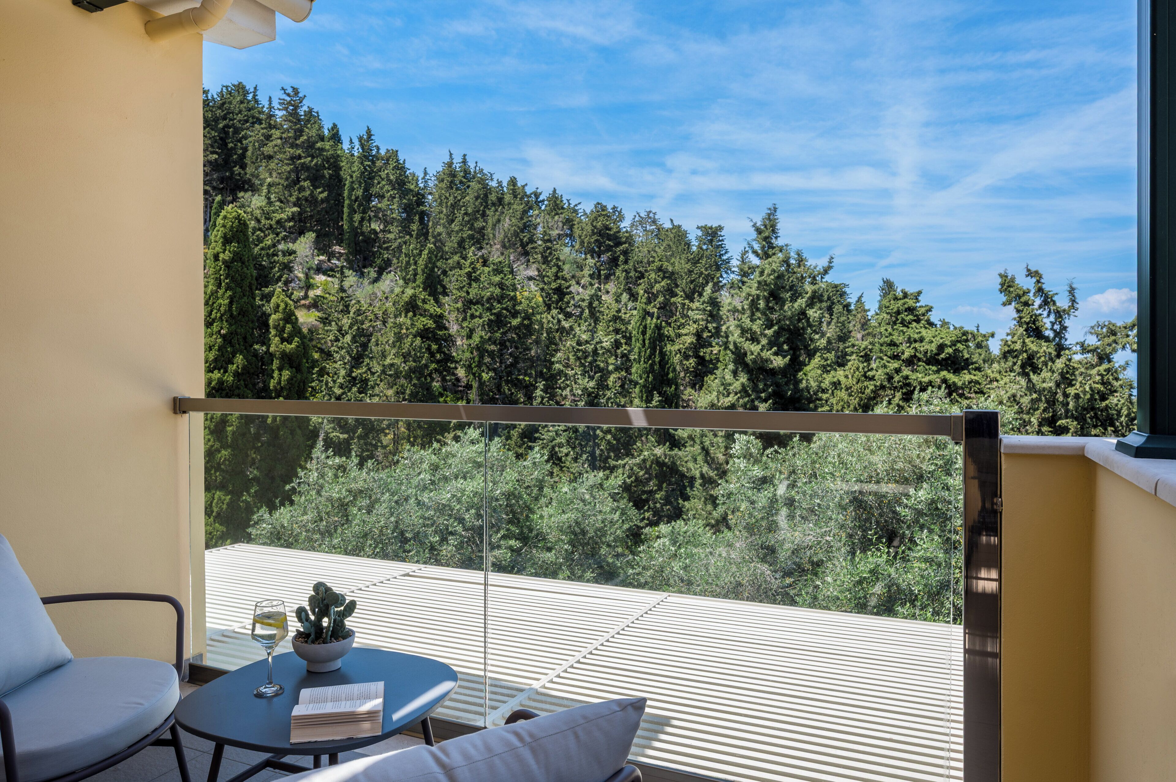A balcony with a view of a forest, a table with a glass of wine, and a chair.