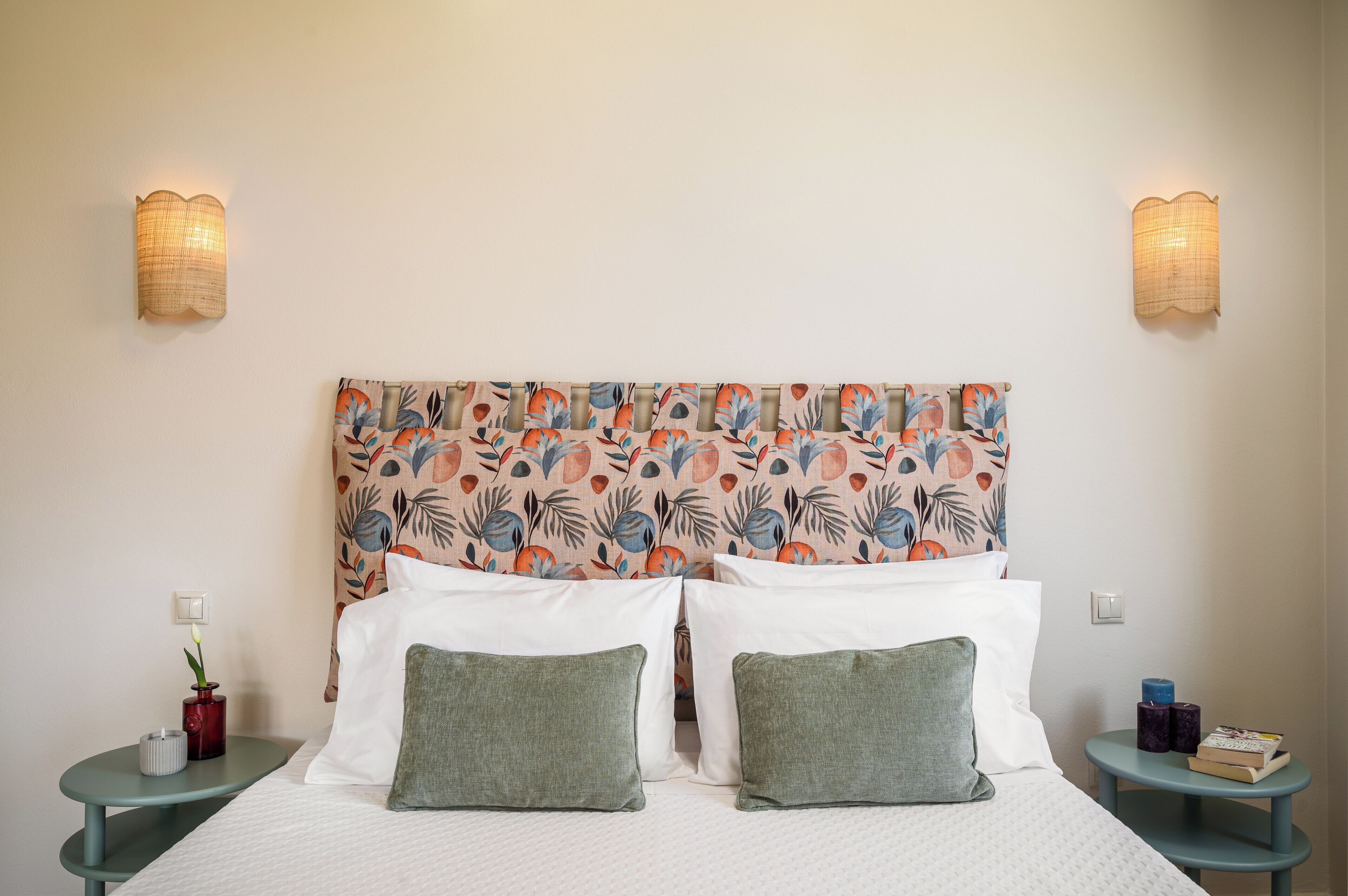 Ein Bett mit weißen Leinentüchern und grünen Kissen, ein Kopfschild mit Blumenmuster, zwei Wandlampen und ein kleiner Tisch mit einer Vase und Kerzen.