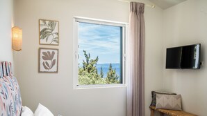 A bedroom with a bed, a TV, a window with a view of trees, and artwork on the wall. - Villas Muscallas by Konnect, 200m from the Beach. (Paxos)