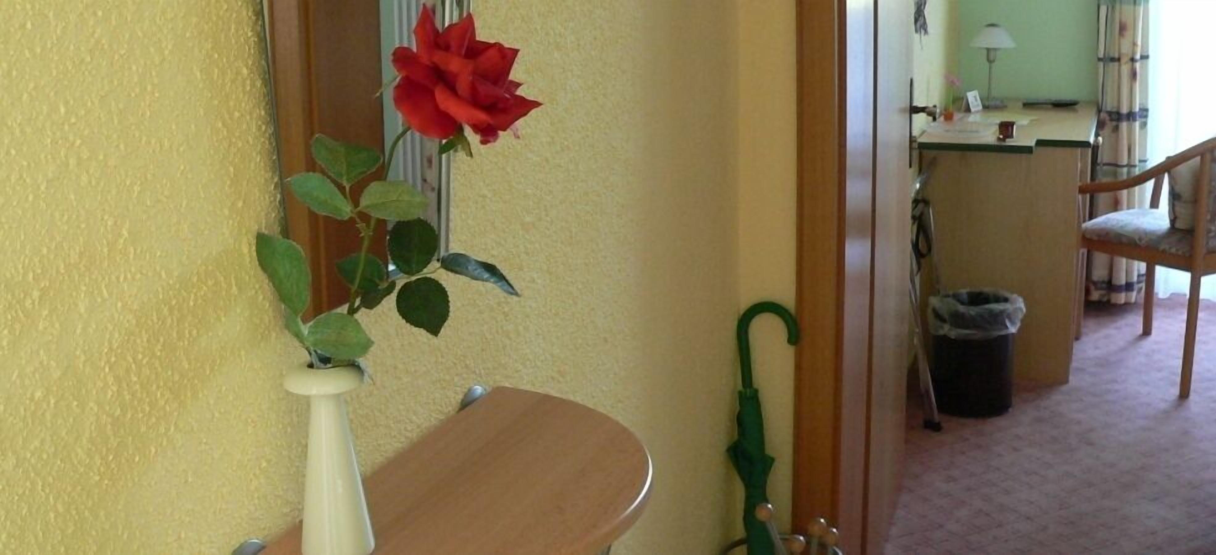 A room with a yellow wall, a wooden chair, a small table, a green umbrella, and a red rose in a vase.