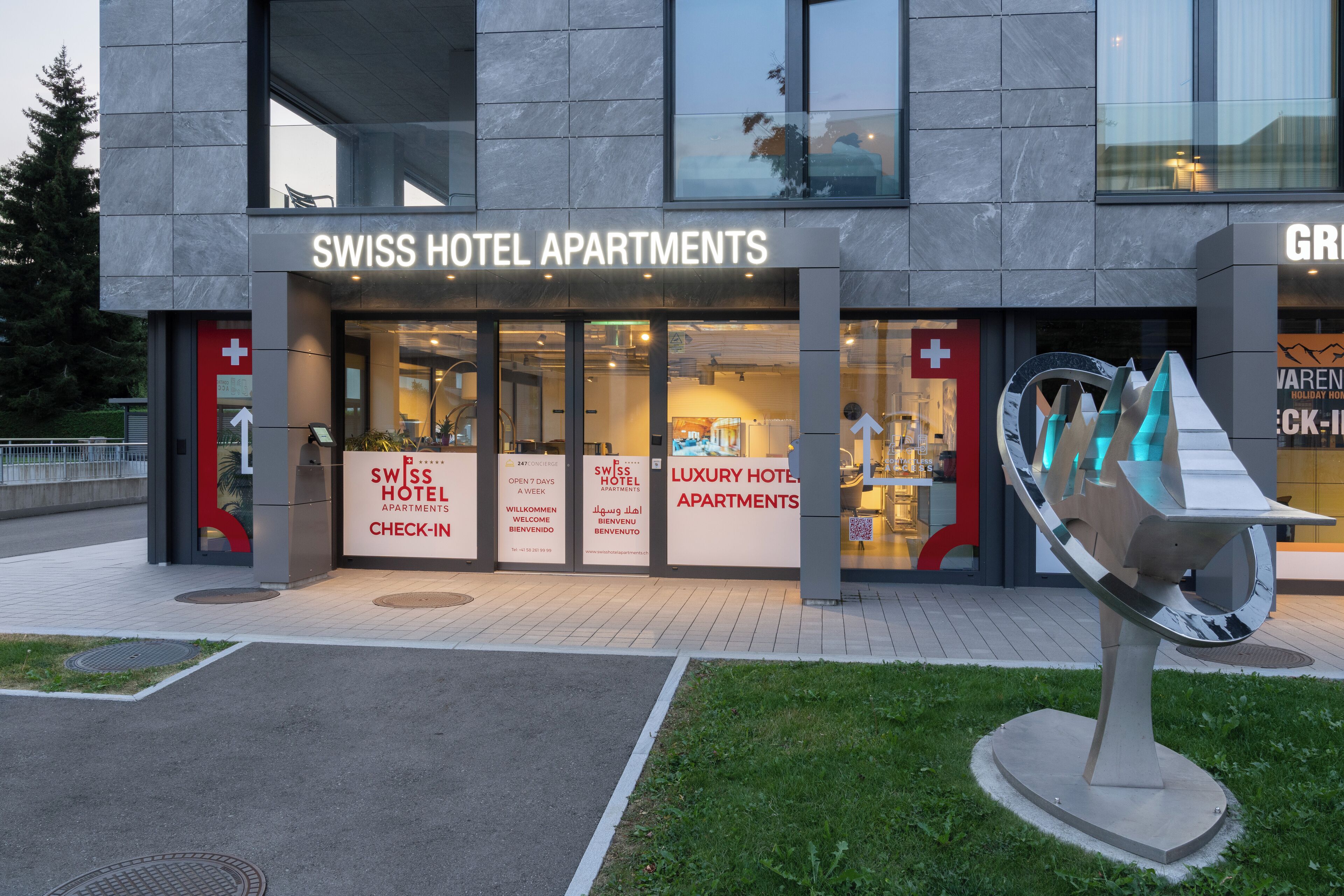 A modern building with a sign for Swiss Hotel Apartments. There are multiple signs in different languages welcoming guests.