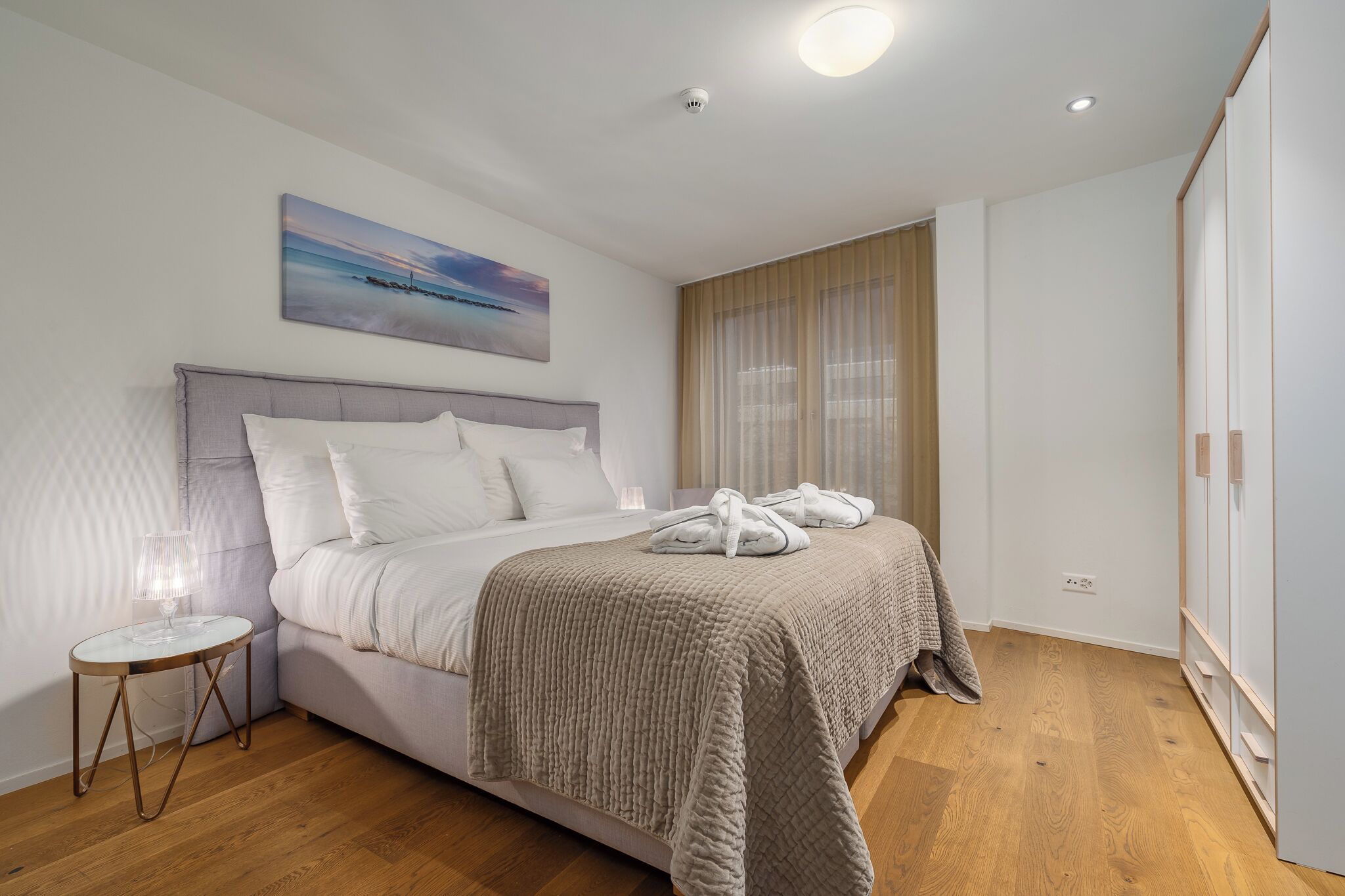 A neatly made bed with a headboard, a bedside table with a lamp, and a painting on the wall.