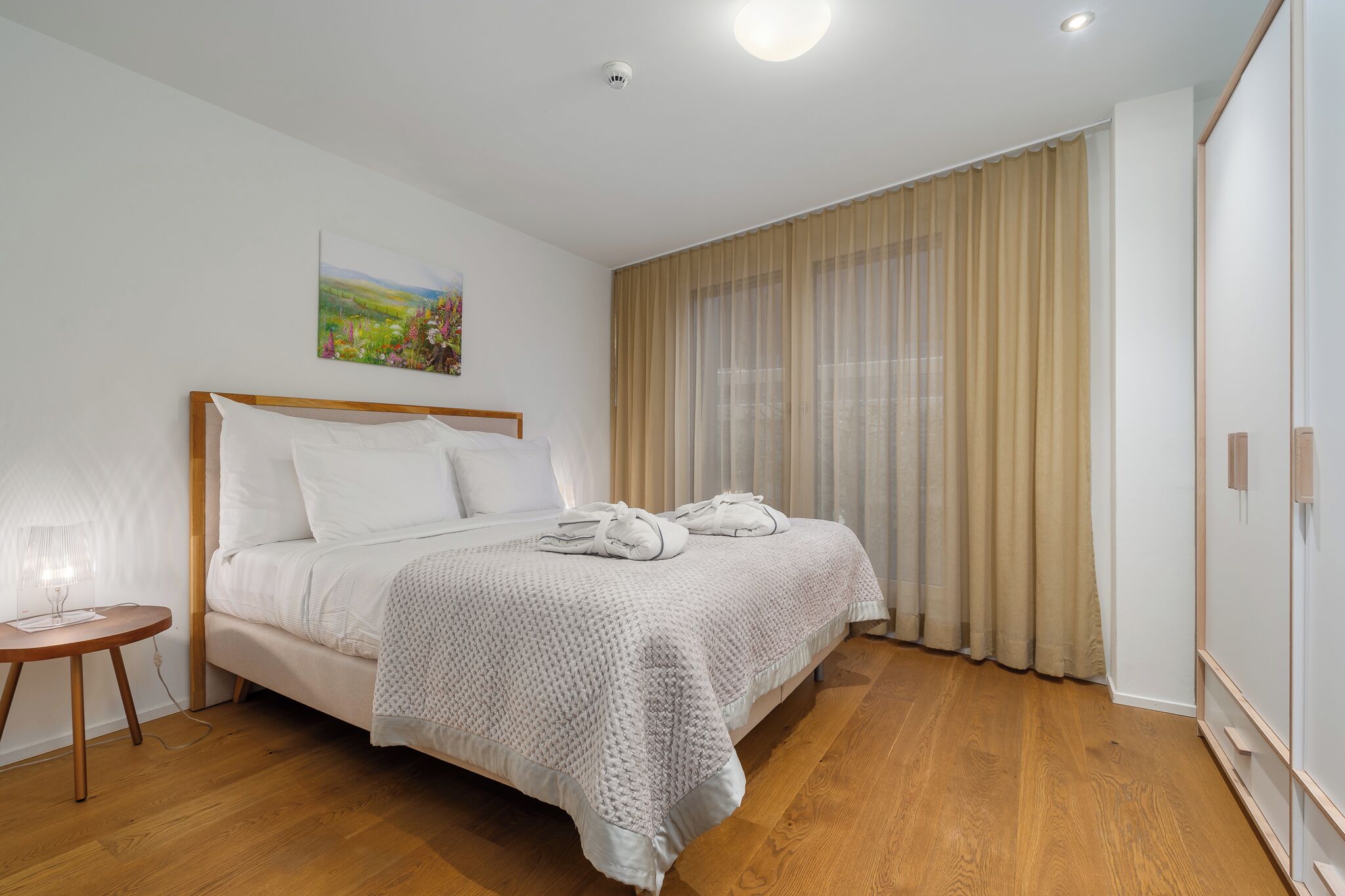 A neatly made bed with a white blanket and pillows, a bedside table with a lamp, and a painting on the wall.