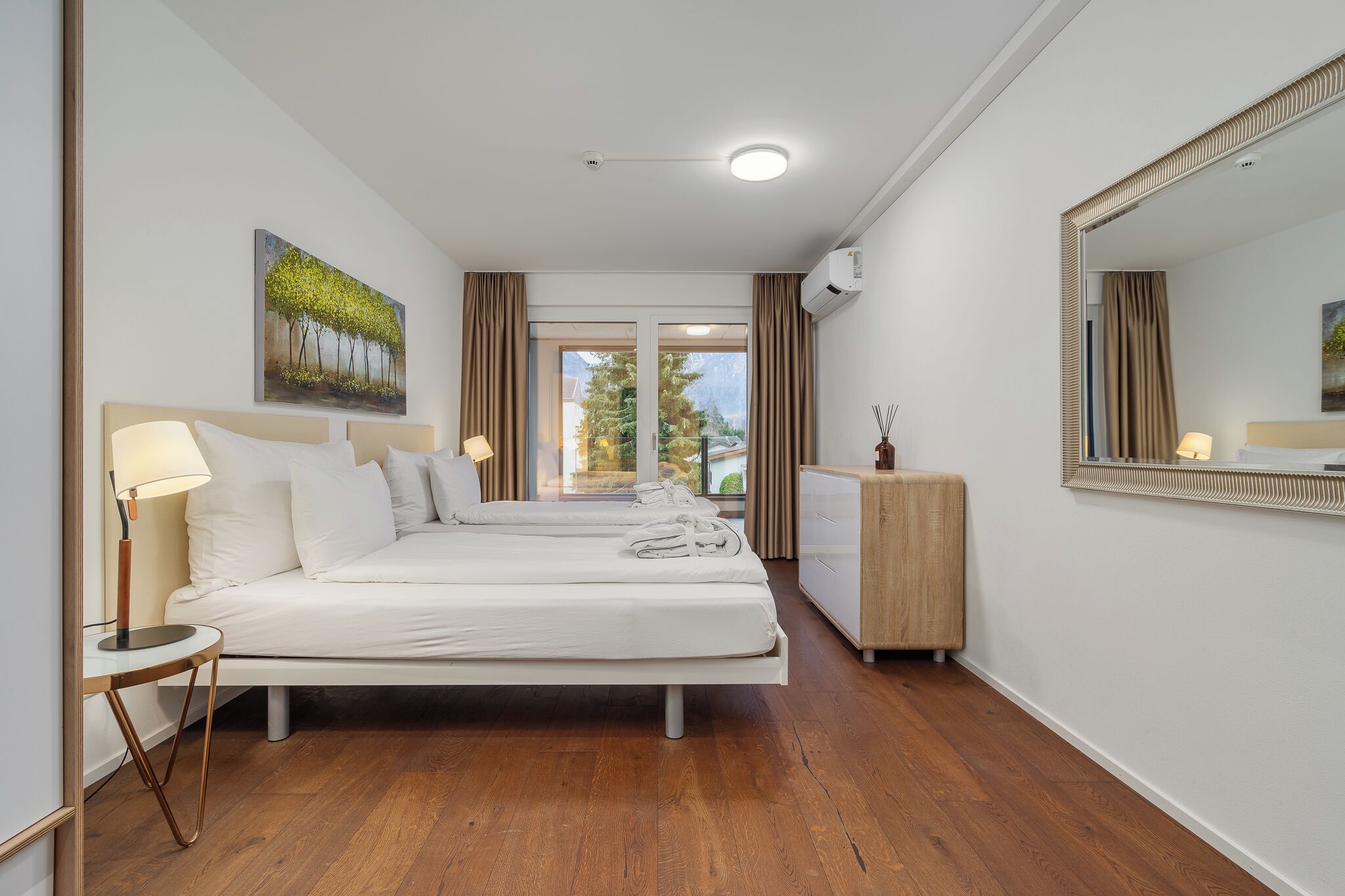 Een hotelkamer met twee bedden, een grote spiegel, een houten vloer en uitzicht op bomen door het raam.
