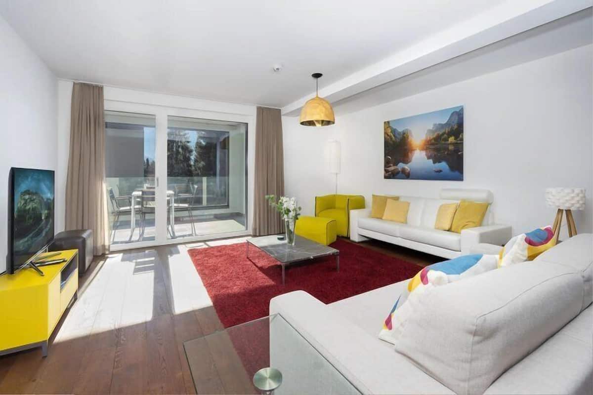 Un salon moderne avec un canapé blanc, un fauteuil jaune et un tapis rouge. Une porte vitrée donne sur un balcon, et un tableau est accroché au mur.