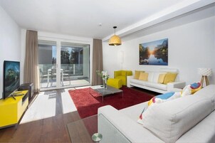 Una sala de estar moderna con un sofá blanco, un sillón amarillo y un tapete rojo. Hay una puerta de cristal que da a un balcón y un cuadro en la pared.