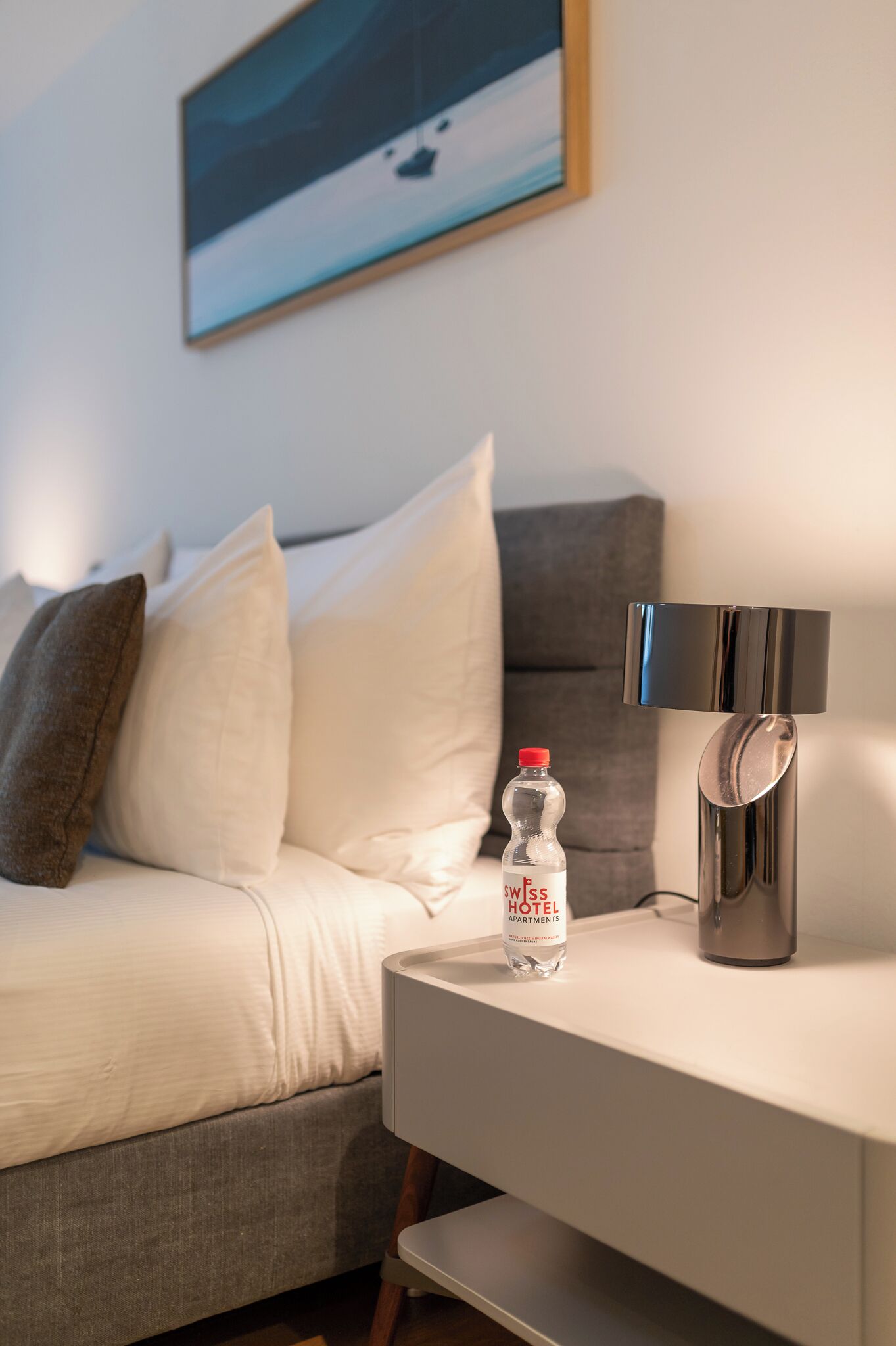 A modern hotel room with a sofa, pillows, a bedside table with a water bottle, a lamp, and a painting on the wall.