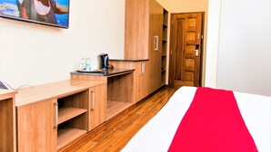 A hotel room with a wooden desk, a bed with a red and white bedspread, a television mounted on the wall, and a wooden wardrobe. - Verona Hotel & Conference Center (Ruiru)