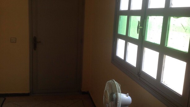 Una habitación con una puerta cerrada, una ventana con fondo verde y un ventilador de pie.