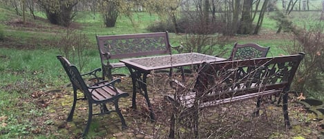 A rustic outdoor seating area with a metal table and benches, surrounded by greenery and trees.