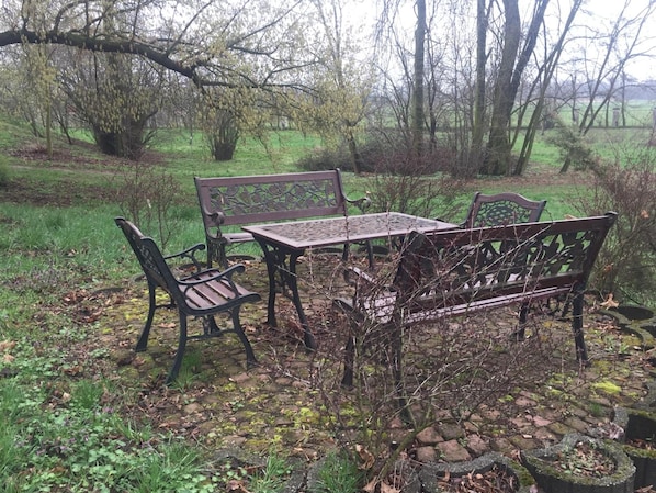 Un espacio de descanso rústico al aire libre con una mesa de metal y bancos, rodeado de follaje y árboles.