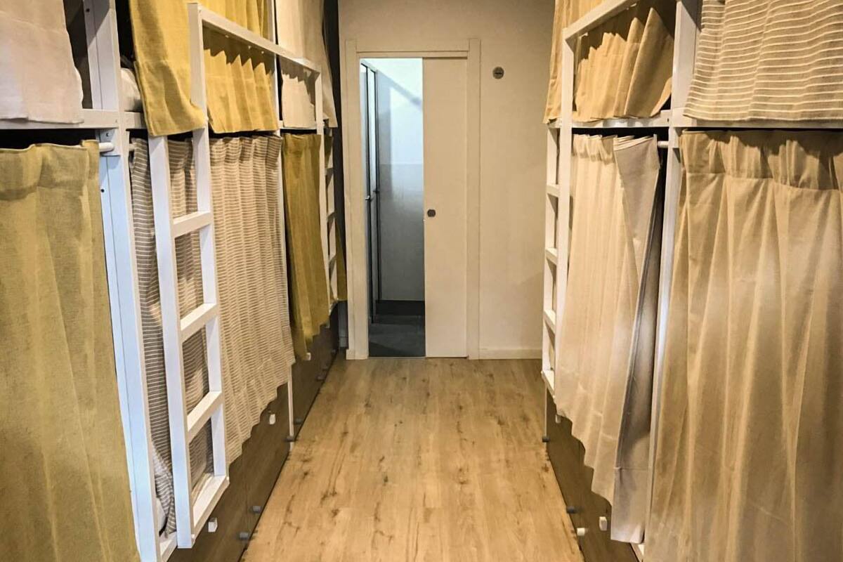 Un pasillo estrecho con suelo de madera, flanqueado por literas de metal blanco con cortinas color beige.