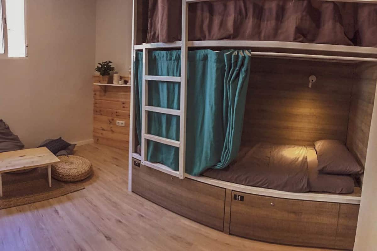 Una habitación con una cama litera, una mesita con cojines, una escalera y un estante con objetos.