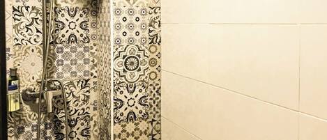 A shower with a patterned tile wall and a plain tiled wall with two towel hooks.