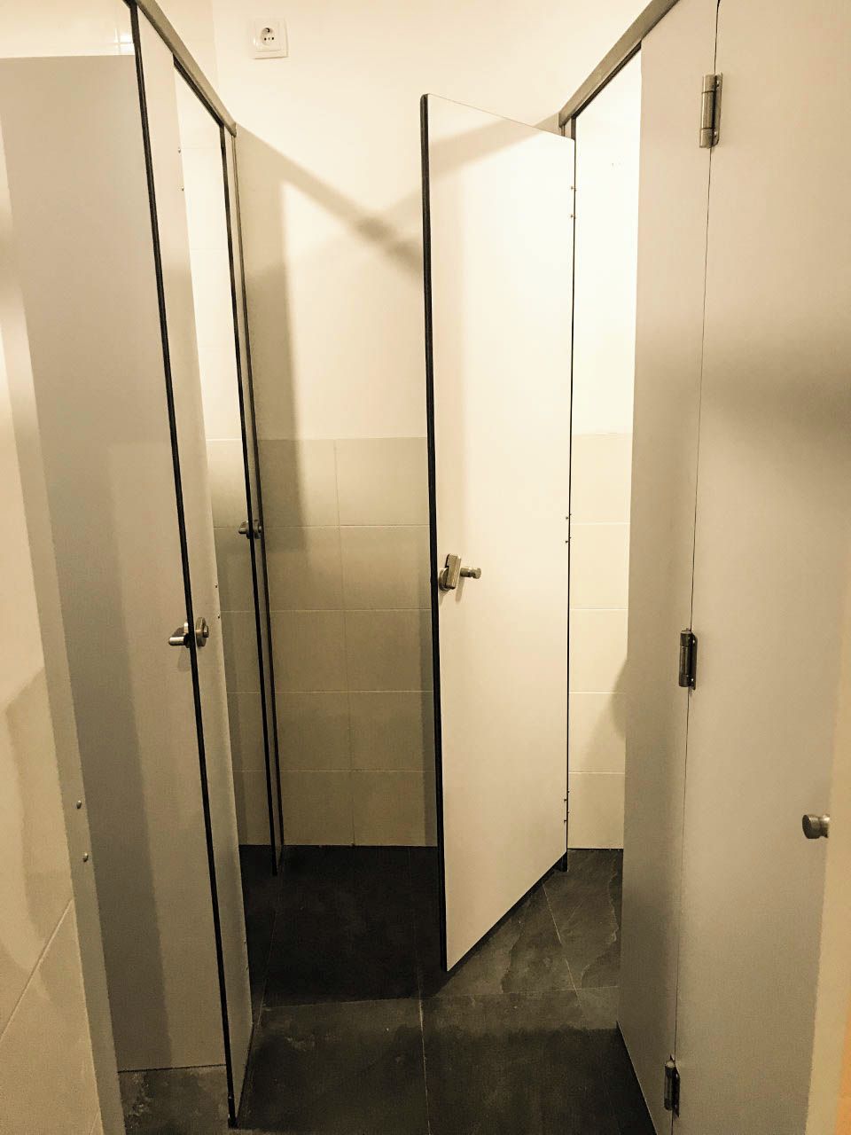 A restroom with three stalls, each with a door and a handle. The floor is tiled, and the walls are plain.