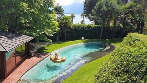 Ein Schwimmbecken mit Holzterrasse, umgeben von üppiger Vegetation und Blick auf ein Gewässer.
