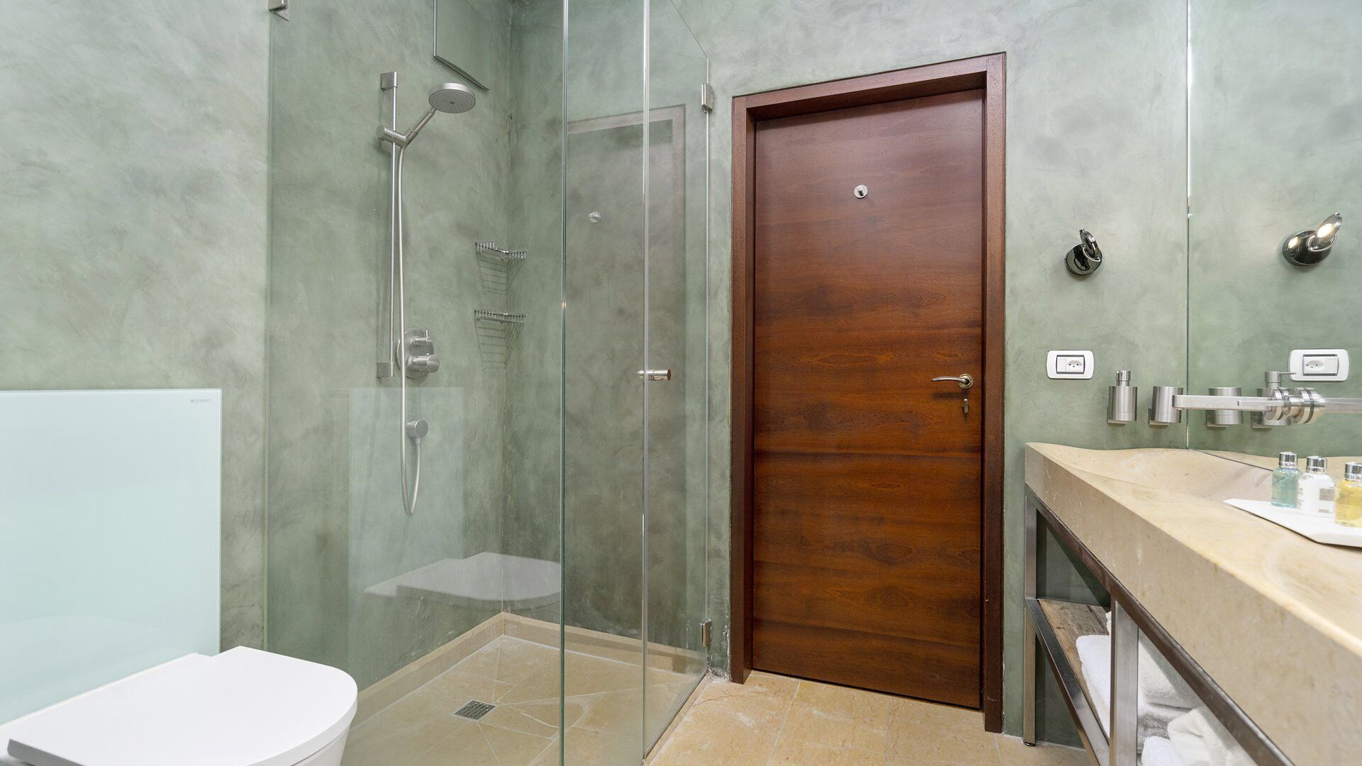 A bathroom with a glass-enclosed shower, a wooden door, a white toilet, and a vanity with a mirror.