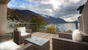 Een balkon met rieten meubilair, een glazen tafel en uitzicht op bergen en een meer.