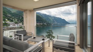 Een balkon met uitzicht op een meer en bergen, ingericht met grijze banken en een tafel.